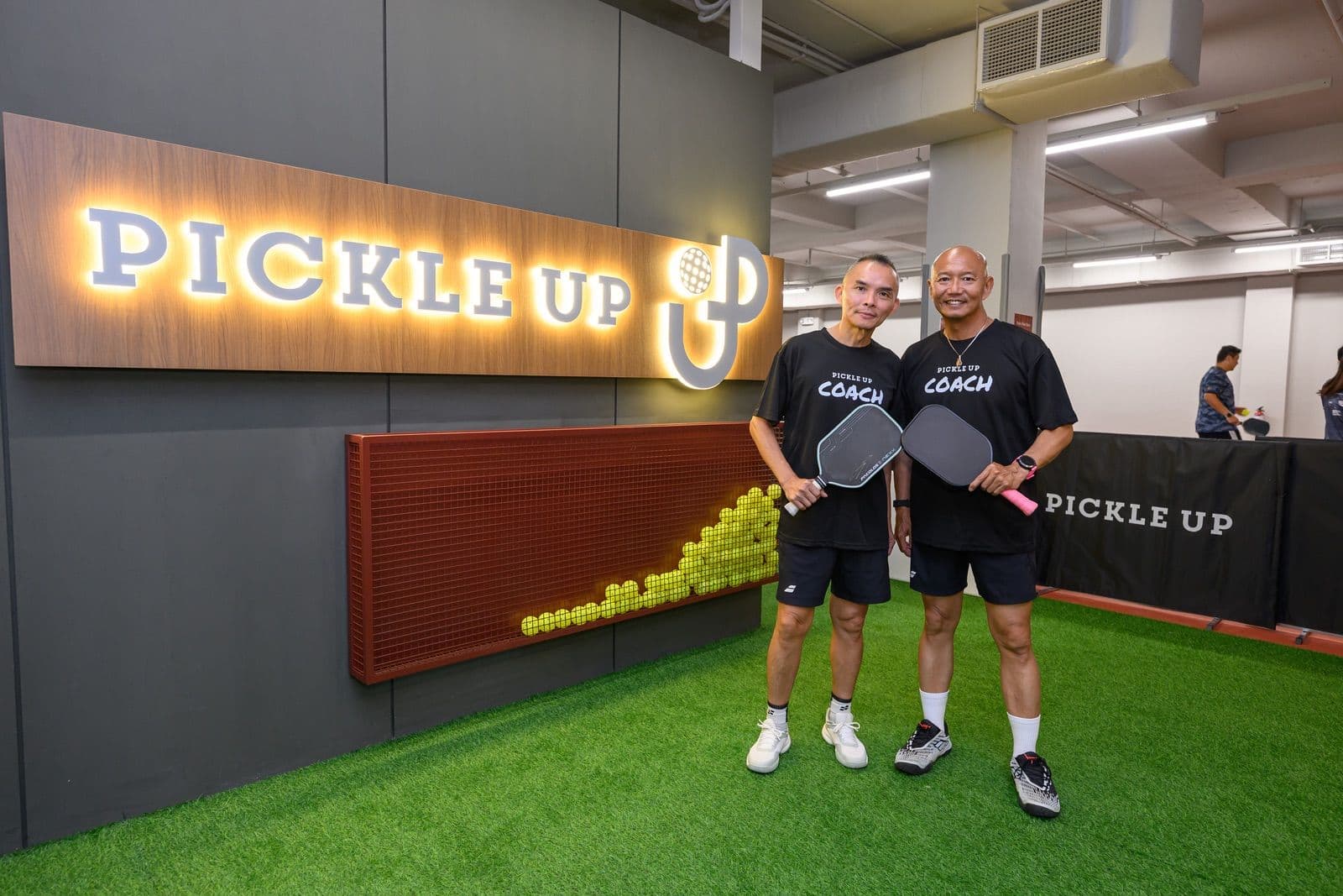 Head Coach SQ and coach Weber stand together in matching Pickle Up Coach shirts by the entrance sign.