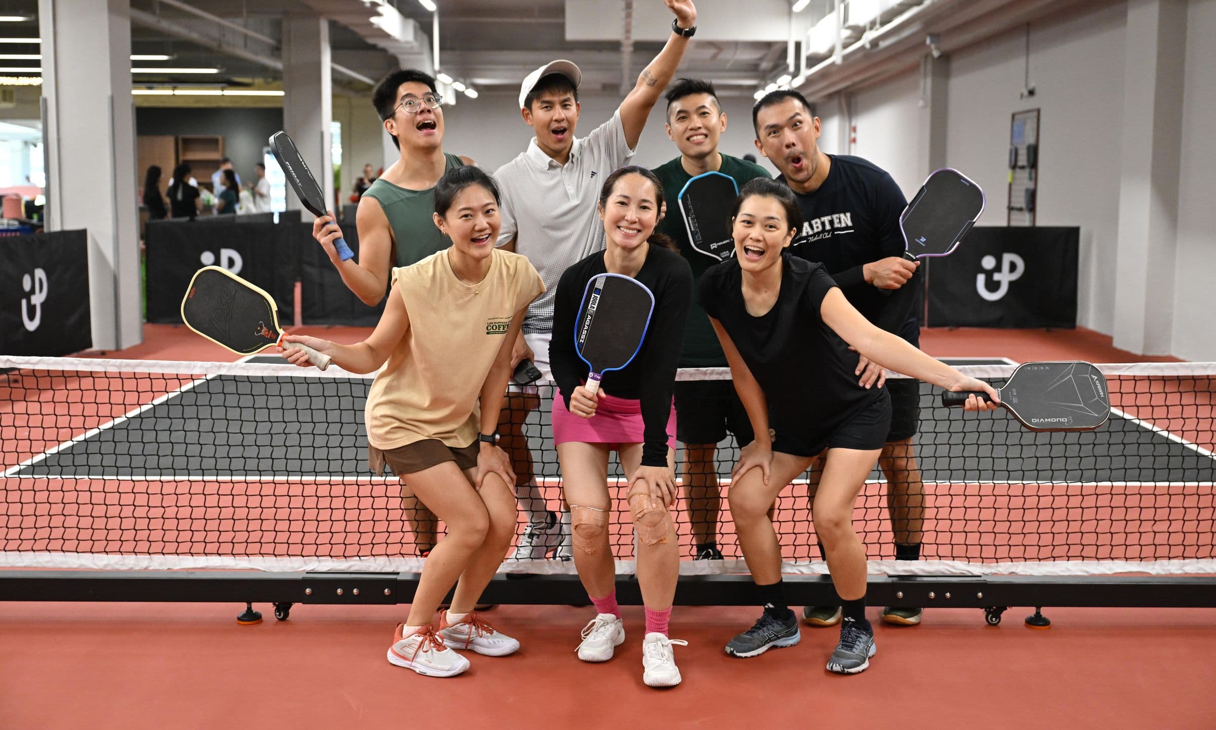 Seven players crowd the net on a Pickle Up court, paddles up and laughing for the camera.