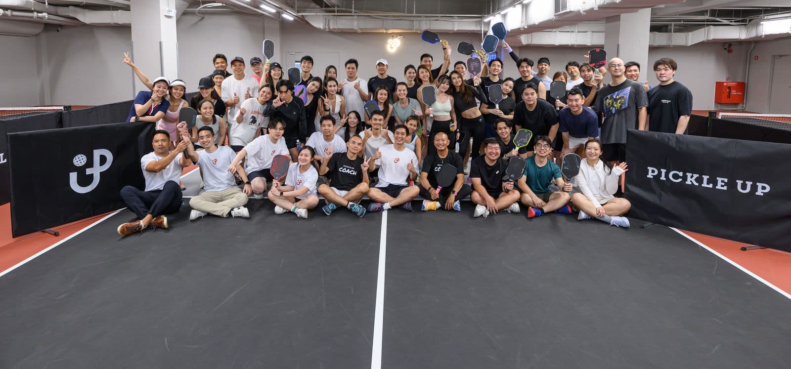 A big group photo on court after a Pickle Up session, everyone smiling and holding paddles up.