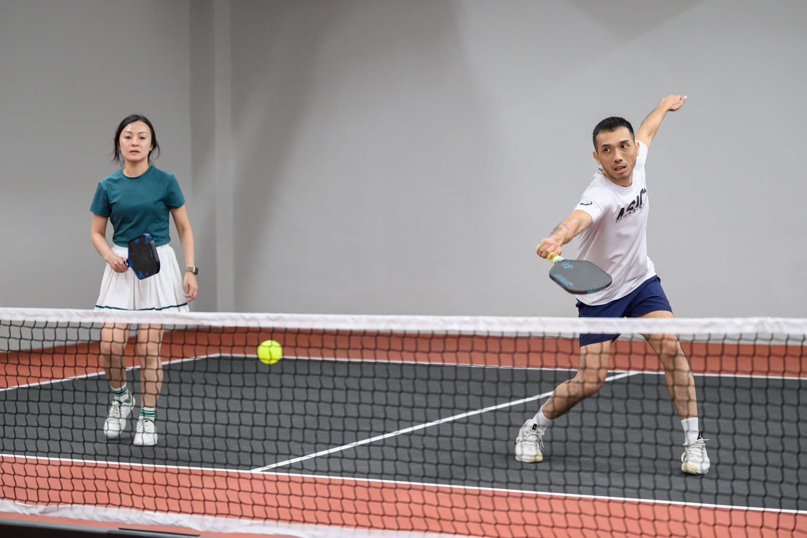 A mixed doubles rally at the net, one player watching the ball, the other swinging through a forehand.